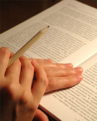 Two hands folded on top of an open book, a pencil intertwined between two fingers.