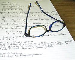 A pair of glasses with thick lenses lying on top of a white writing tablet on which are written some notes.