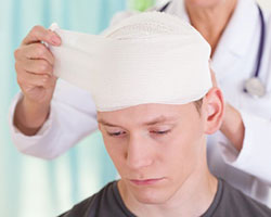 A downcast-looking young man, whose head is being bandaged by a medical professional who is standing behind him.