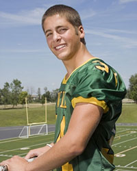 A healthy looking Josh, in a football uniform, is smiling into the camera while standing on a balcony overlooking a green football field.