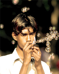 A young man facing the viewer at an angle, staring distractedly and creating a stream of bubbles by blowing through a plastic device.