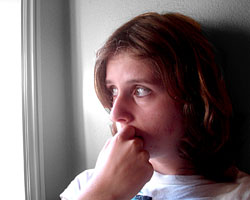 A close-up of a young woman in semi-profile with her hand at her mouth, looking out a window apprehensively.