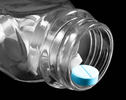 A see-through pill bottle, on its side with no cap, white pills inside, and a blue tablet on the rim awaiting pickup.
