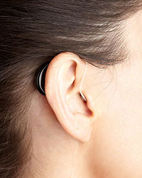 A close-up, side on, of an ear with an almost unnoticeable hearing assistance device that hooks over the ear flap. The device has a thin, see-through plastic line that leads to the hearing aid inside the ear canal.