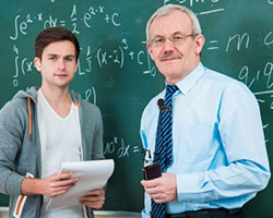 A young male student holding a note pad is standing next to an older male professor. Both are facing the camera. Behind them is a green blackboard containing math formulas written in white chalk.