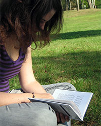 A young woman sitting cross legged on the grass near a tree which is partially shading her upper body and head. She is looking down, poring over an open book, her face obscured by her long, dark hair.