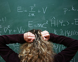 A view of the back of a person’s head with long, wavy black hair with blond highlights. The person is facing a blackboard filled with mathematical formulas. He or she is pulling at his or her hair.