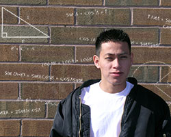 A young man with a white T-shirt and open black leather jacket, facing the viewer. Behind him is a red brick wall covered with algebraic symbols and equations drawn in white chalk.