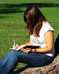 A young woman with long, dark hair sitting on a campus lawn at the base of a tree, writing in a notebook with a large pen.