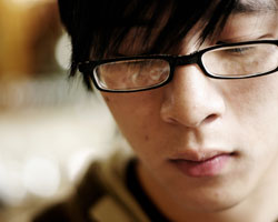 A young man’s face, facing the viewer, black hair, head tilted downwards, wearing black plastic glasses that obscure his eyes, the lenses of which have reflections in them.