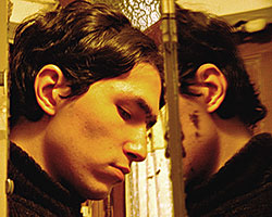 A head shot of a young man with dark, unkempt hair, in profile, looking downcast, in front of a mirror in which he is partly reflected.